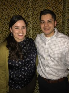 A woman and a man are smiling while standing side by side in front of a patterned wall. The woman has long dark hair and is wearing a dark patterned top and a green cardigan. The man is wearing a plaid shirt.