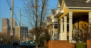 A residential street with trees and parked cars, featuring traditional houses with porches on the right. In the background, city skyscrapers are visible under a clear blue sky.