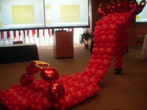 A display of red balloons forms a pathway, leading upwards in a wave-like pattern in a conference room. Large screens and flags in the background indicate a formal setting.