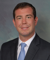 A man in a suit and tie poses against a neutral background. He has short brown hair and a slight smile.