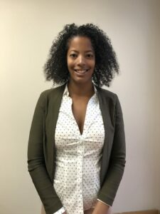 A person with curly hair smiles at the camera. They are wearing a green cardigan over a white shirt with black polka dots. The background is a plain, light-colored wall.