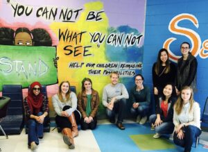 A group of nine people pose in front of a colorful mural. The mural features a girl holding a sign that reads "Stand" and includes the text, "You cannot be what you cannot see." The setting is a brightly painted room.
