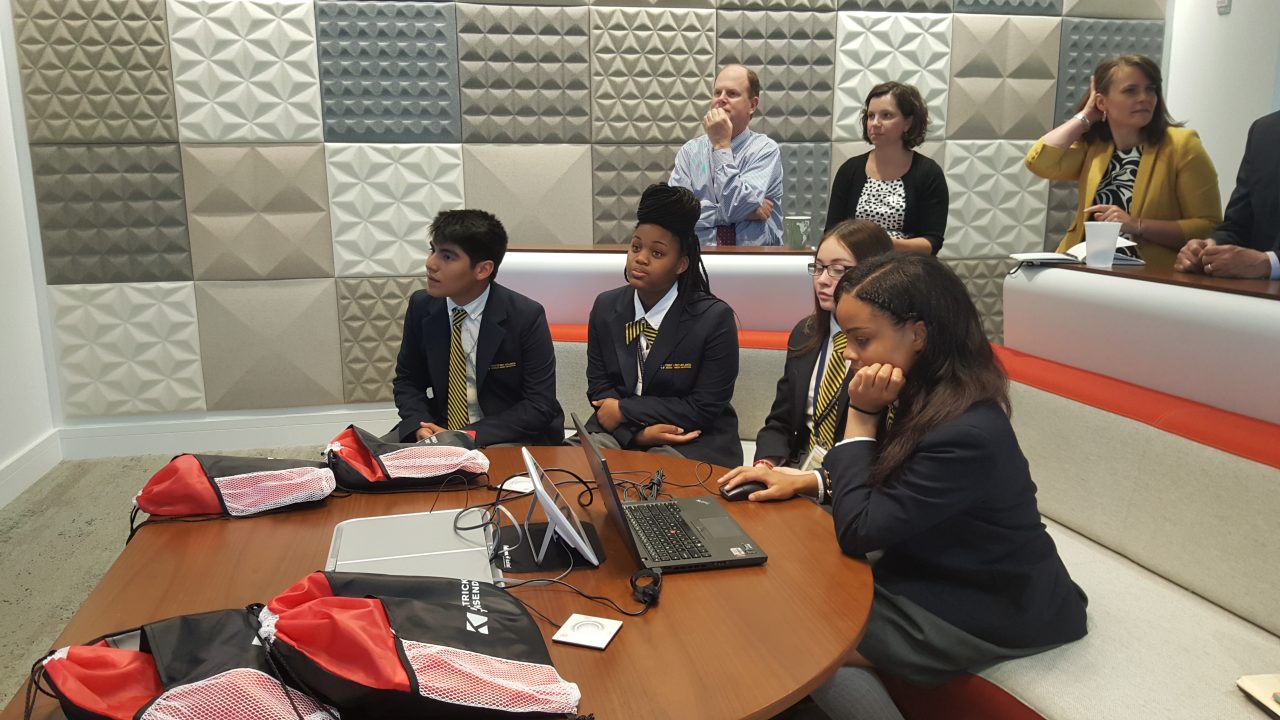 A group of students in uniform sit at a table with laptops and backpacks, engaged in a presentation. Adults stand behind, observing. The room has a modern design with patterned acoustic panels on the wall.