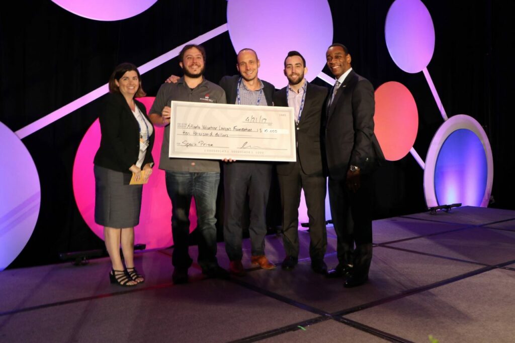 Five people are standing on a stage holding a large check. The background features circular designs with pink and purple lighting. They appear to be celebrating or presenting the check.