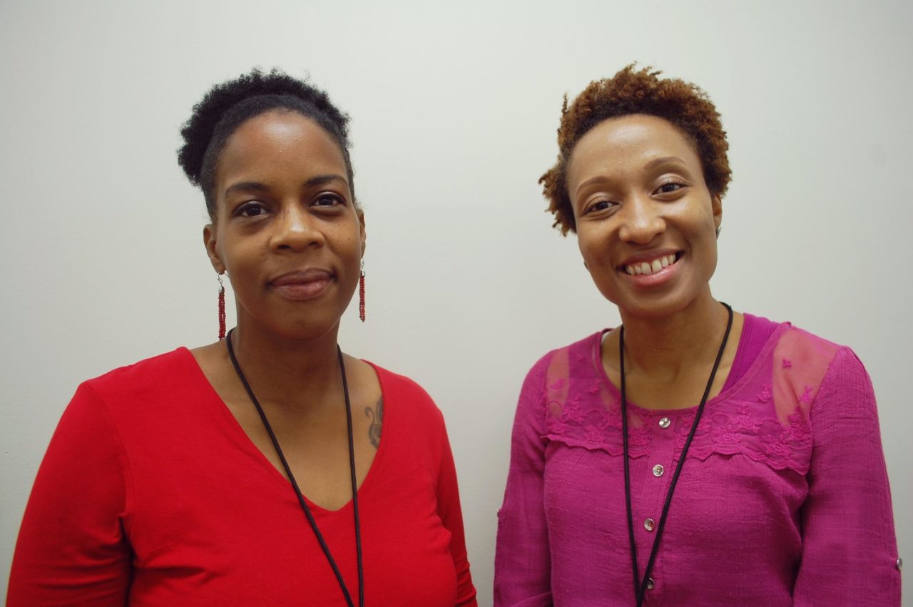 Two people are standing in front of a light-colored wall, smiling. One is wearing a red top with earrings, and the other is in a magenta top with lace details. Both have name badges on lanyards around their necks.