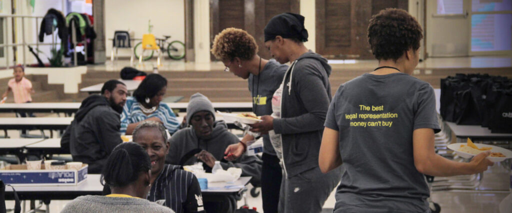 People are seated at tables in a cafeteria setting, eating and conversing. Two people wearing shirts with text on the back are serving food. The text reads, "The best legal representation money can't buy.