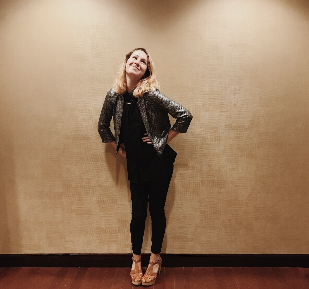 A person with shoulder-length hair poses confidently against a beige wall. They wear a shiny jacket, black shirt and pants, and brown shoes, smiling with hands on hips.