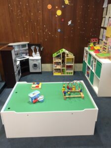 A children's play corner with a play kitchen, colorful toy storage shelves, a small playhouse, an activity table with an ice cream play set, and a bead maze. The wooden wall is decorated with a space-themed mural and kids' drawings.