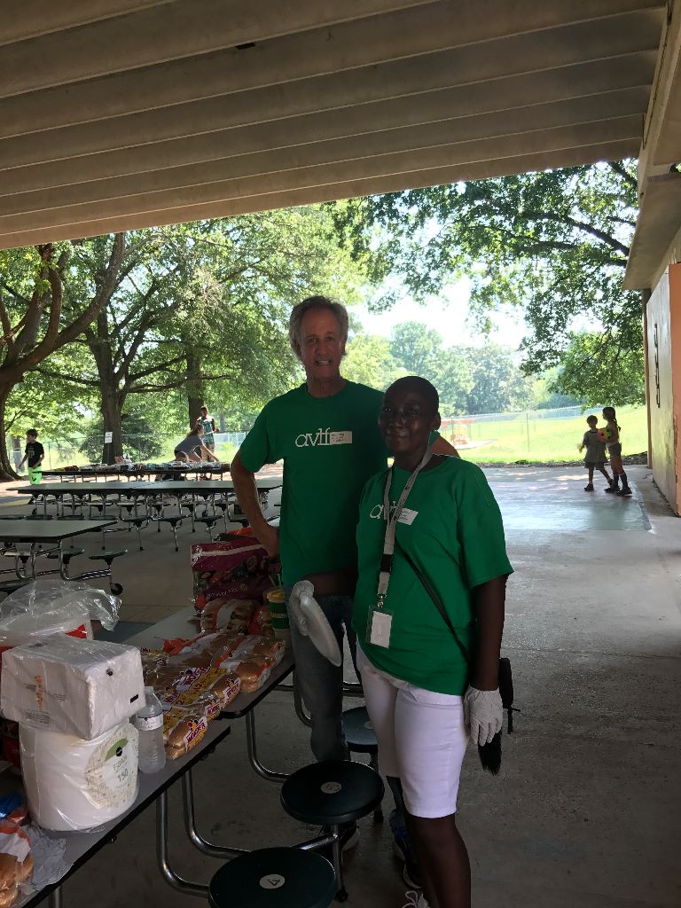 Two people in green shirts stand behind a table with supplies, including paper towels and food items, in an outdoor pavilion with picnic tables. Trees and a grassy field are visible in the background.