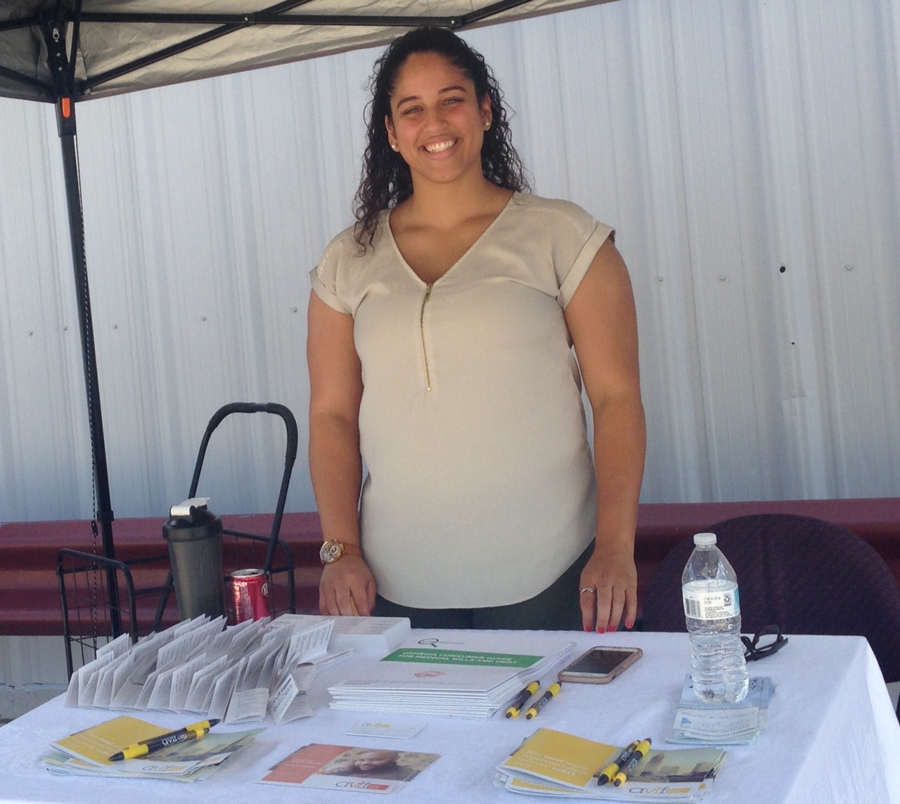 A woman stands behind a table covered with flyers, pamphlets, pens, and a water bottle. She is smiling, under a canopy, with a corrugated metal wall in the background.