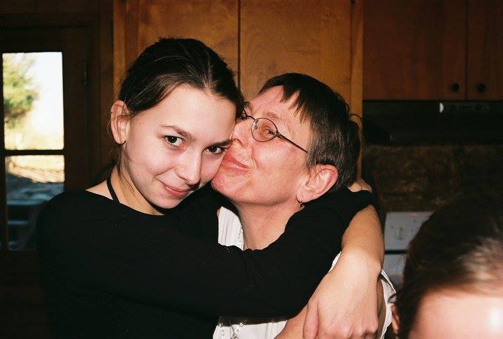 Two people are hugging in a warmly lit room. The person on the left has dark hair and is smiling at the camera, while the person on the right, wearing glasses, is kissing the other's cheek affectionately. A window and wooden cabinets are in the background.