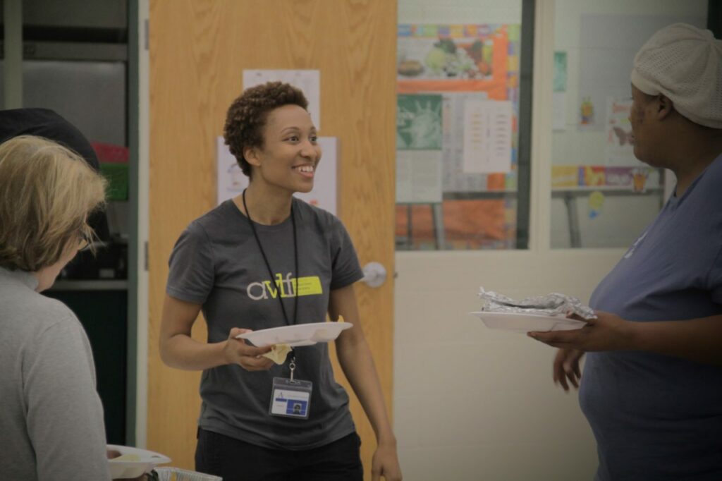 A smiling person wearing a gray t-shirt holds a plate of food, engaging with another person in a blue shirt who is also holding a plate. They are in a room with educational posters on the walls.