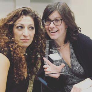 Two women are sitting together. One is smiling broadly and holding a transparent award. The other has long curly hair and is looking to the side. They appear to be in a celebratory setting indoors.