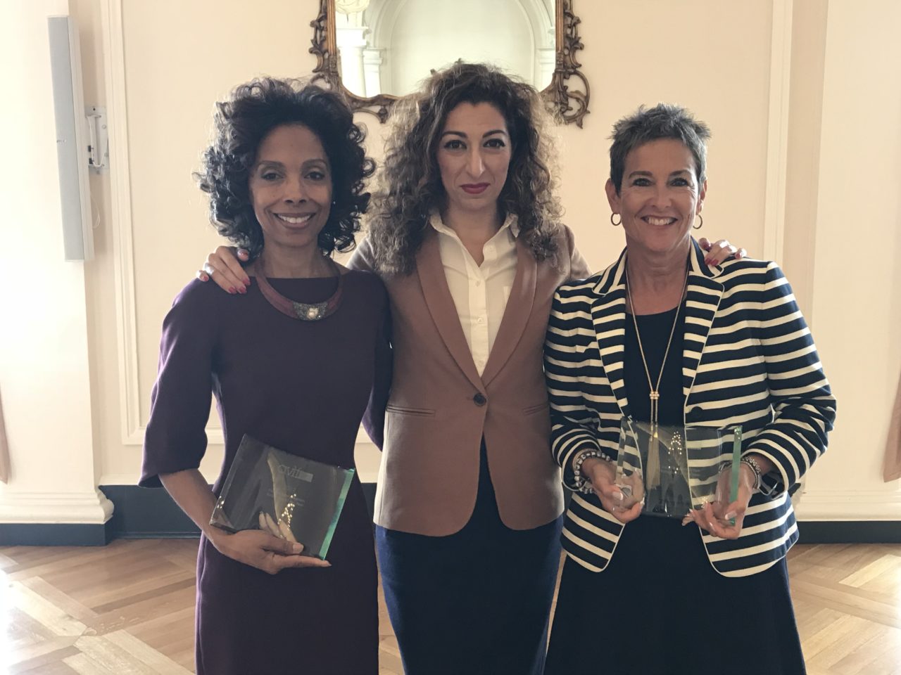 Three women stand together, smiling, holding awards. The woman on the left wears a purple dress, the middle woman is in a tan blazer, and the woman on the right wears a striped blazer. They are in a room with wooden floors and light-colored walls.