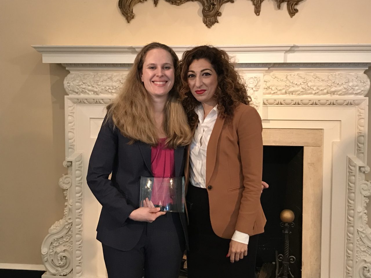 Two women standing in front of an ornate fireplace. One woman, wearing a dark suit and pink blouse, is holding a glass award and smiling. The other woman, in a tan blazer and white blouse, is smiling with her arm around the award recipient.