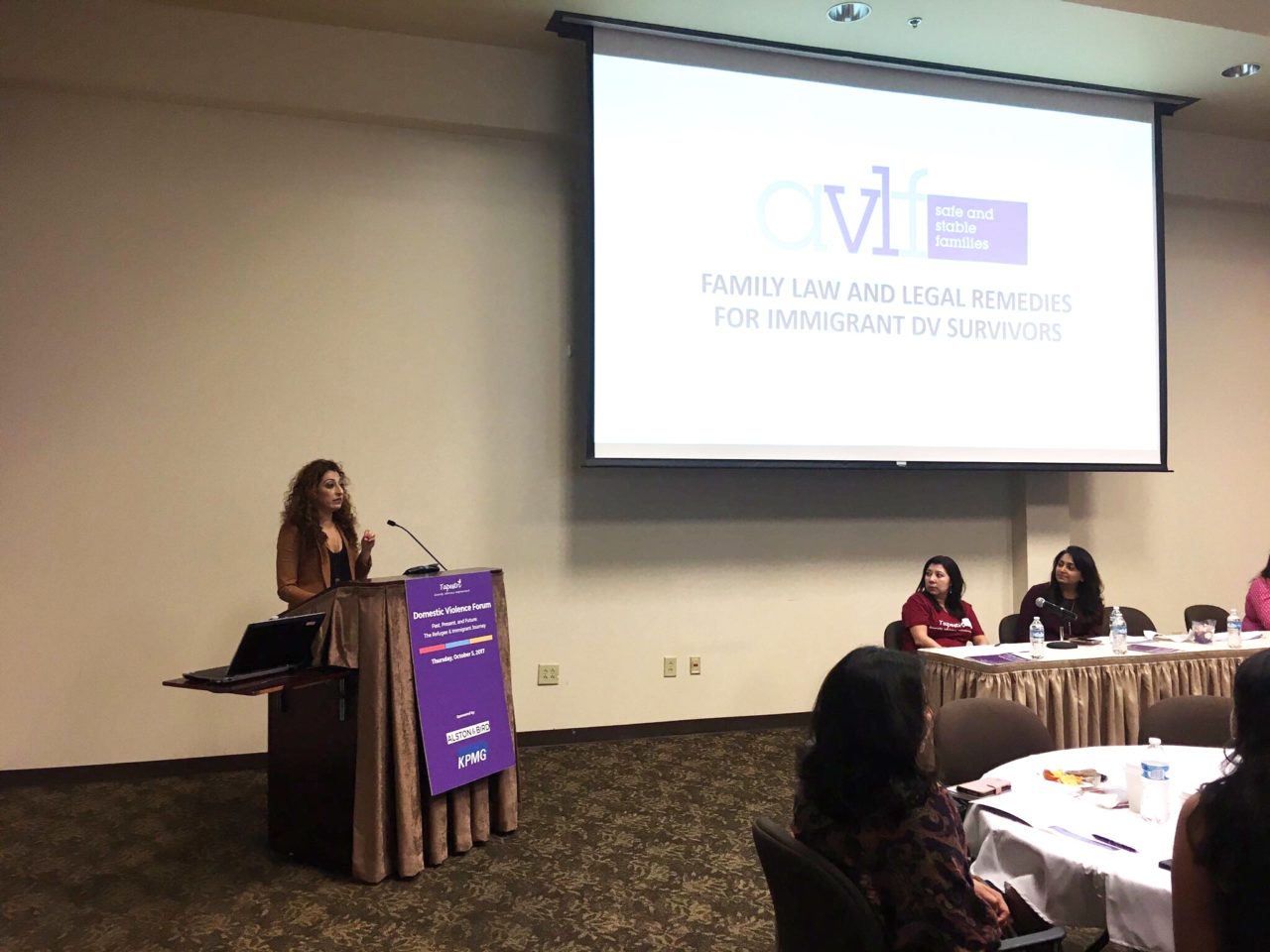 A woman stands at a podium giving a presentation, with a screen displaying "Family Law and Legal Remedies for Immigrant DV Survivors." Several people are seated at a table listening.