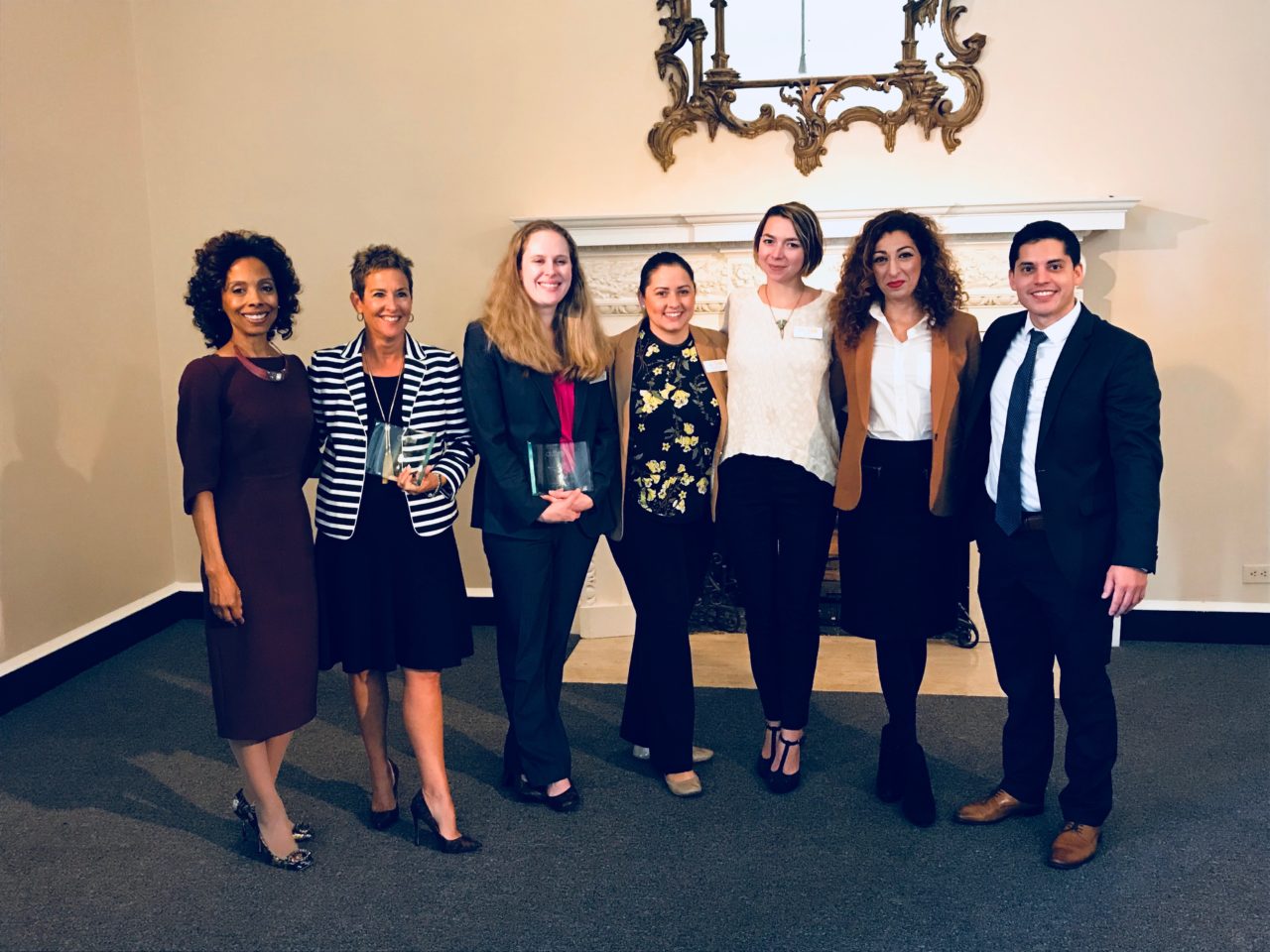 A group of seven people, dressed in business attire, stand together in a well-lit room. Two individuals hold glass awards. A decorative mirror and fireplace are in the background.