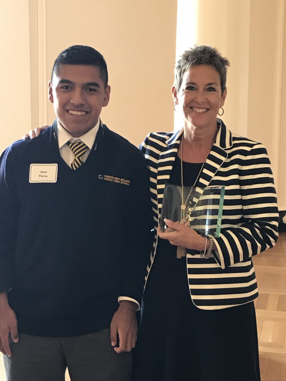 A young man in a uniform stands next to a woman holding an award. Both are smiling and standing in a warmly lit room. The woman is wearing a striped jacket, and the young man has a name tag.