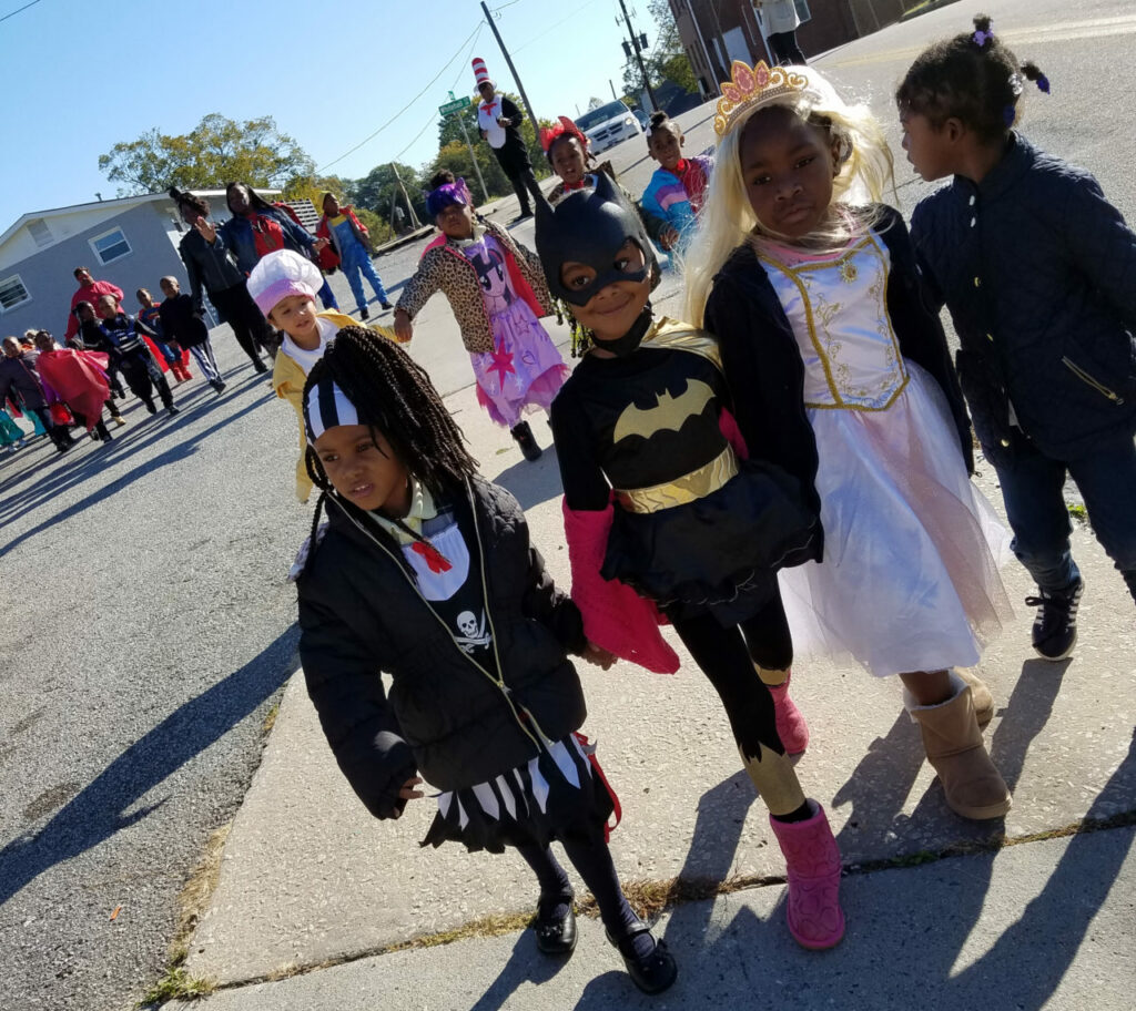 A group of children in Halloween costumes walk along a street. One wears a bat costume, another is in a princess outfit, and a third has a skeleton theme. Other children in colorful costumes follow them, with adults nearby. The sky is clear and sunny.