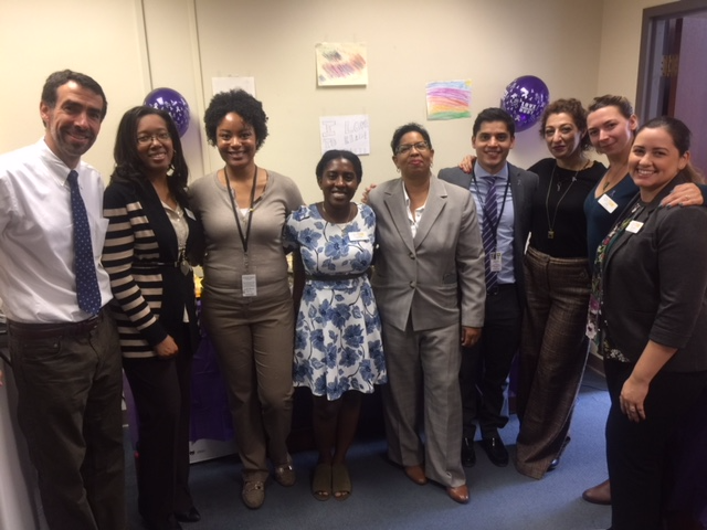 A group of nine people stands in a room with beige walls, smiling at the camera. The room has colorful drawings and purple balloons on the walls. Everyone is dressed in business casual attire.