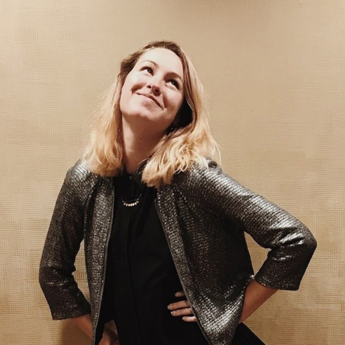 A person with shoulder-length hair stands against a plain background, wearing a metallic jacket over a dark top, looking upwards with a slight smile and hands on hips.