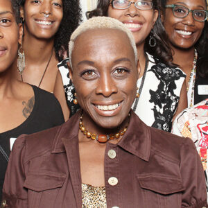 A group of women smiling at the camera. The woman in front has short blonde hair, wearing a brown jacket and a beaded necklace. Others in the background are smiling warmly.