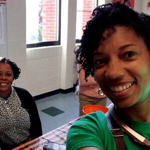 Two people smiling in a brightly lit room with windows. One takes a selfie wearing a green shirt, while the other stands in the background near a table covered with an orange and black cloth.