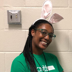 Person wearing a green shirt and glasses, smiling, with pink bunny ears headband, standing against a light-colored wall.