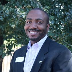 A person in a dark suit and light-colored shirt smiles while standing in front of a tree with lush green leaves. The background is a mix of foliage and blurred light.