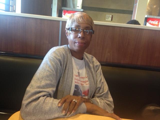 A person with short blonde hair and glasses is sitting at a table in a restaurant. They are wearing a gray jacket and a white t-shirt with red text. The restaurant has wooden panels and beverage signs in the background. The image is upside down.