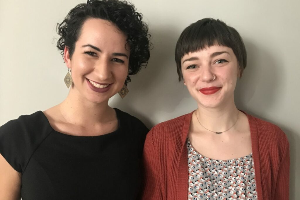 Two smiling women stand side by side against a light gray background. The woman on the left has curly dark hair and is wearing a black top and dangling earrings. The woman on the right has short hair and is wearing a red cardigan over a patterned blouse.