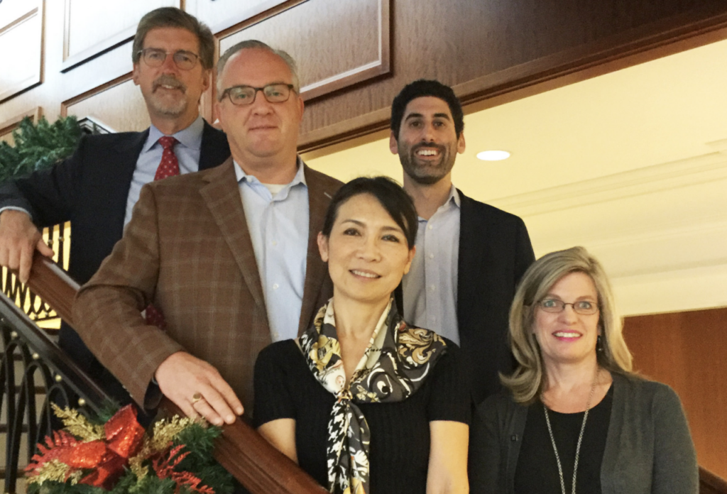 A group of five people professionally dressed, standing and smiling on a staircase indoors. The rail is decorated with festive greenery and ribbons. They are positioned at different levels of the stairs, with wood paneling in the background.