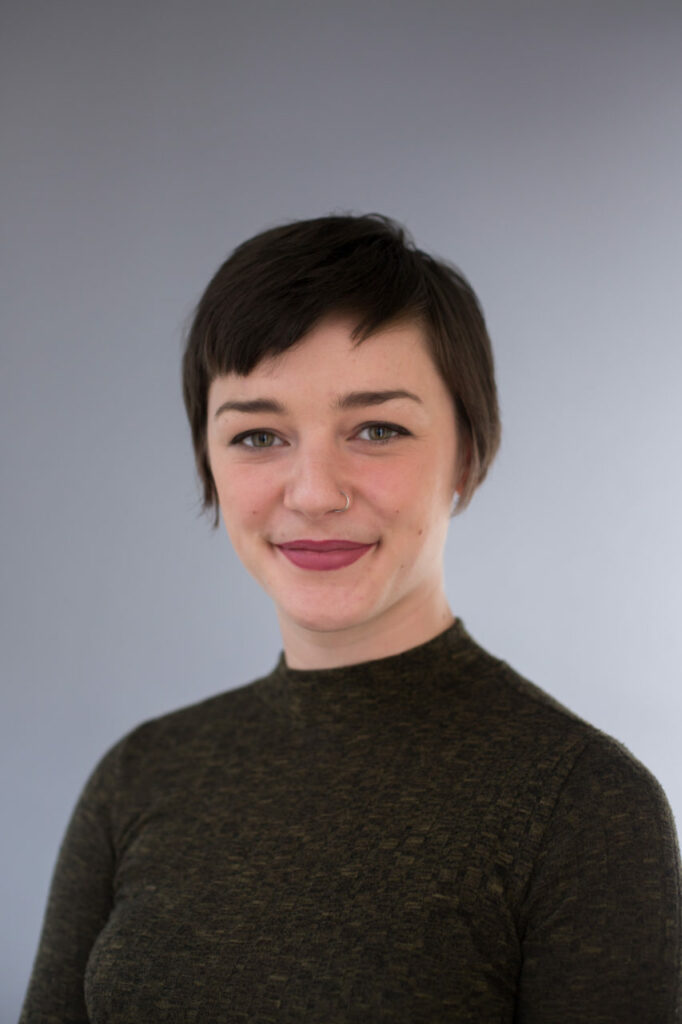 A person with short dark hair is smiling at the camera. They are wearing a dark green long-sleeve top and have a nose ring. The background is a plain grey color.