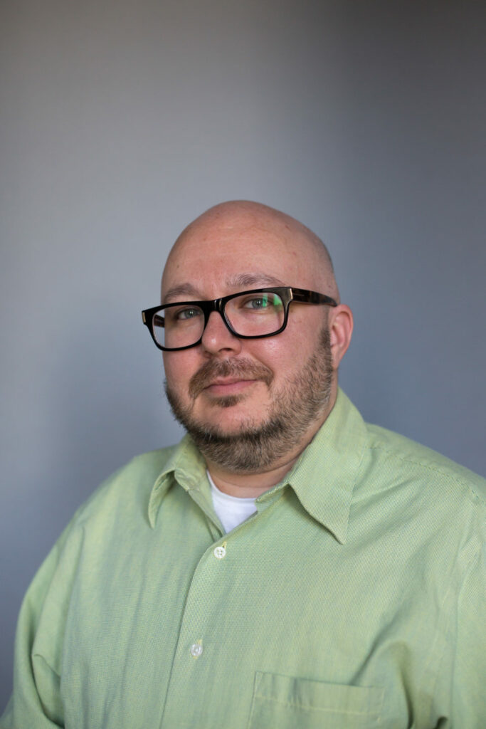 A man with glasses and a beard is wearing a light green shirt. He is looking at the camera with a neutral expression. The background is a plain gray color.