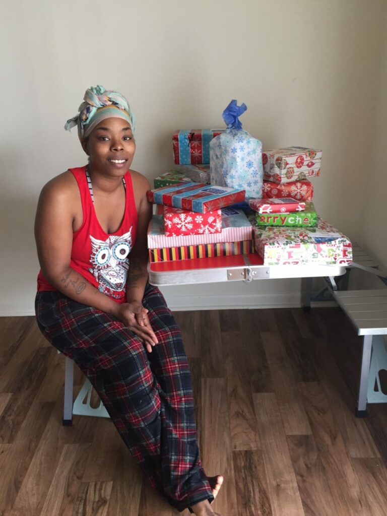 A woman in a red tank top and plaid pajama pants sits on a bench next to a table filled with various wrapped gifts. She smiles at the camera, and the gifts are stacked in colorful wrapping paper.