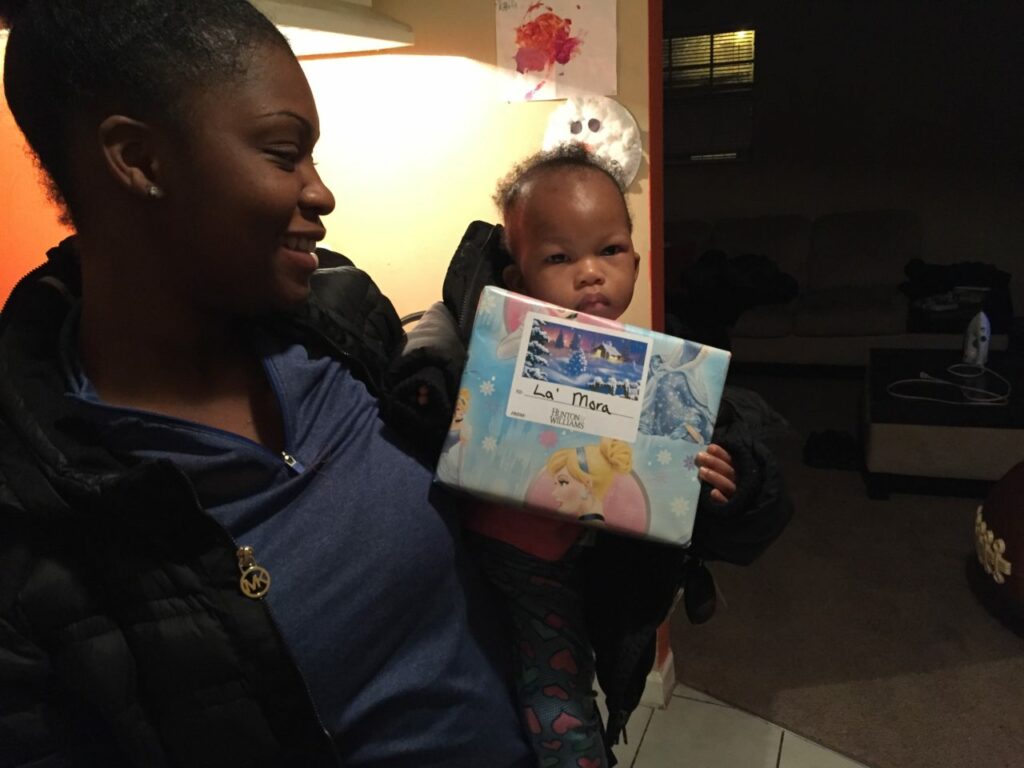 A woman holding a baby who is holding a gift wrapped in Frozen-themed paper. The woman is smiling at the baby. A dimly lit room with a couch and wall art is in the background.