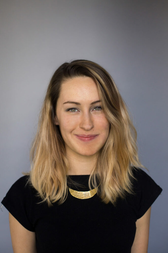 A person with shoulder-length blonde hair and a slight smile is wearing a black top and a golden necklace. The background is a plain gradient of gray.