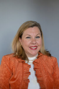 Woman smiling in a professional portrait; she has shoulder-length blonde hair and is wearing an orange jacket over a white blouse, accessorized with pearl earrings and a necklace. The background is plain gray.