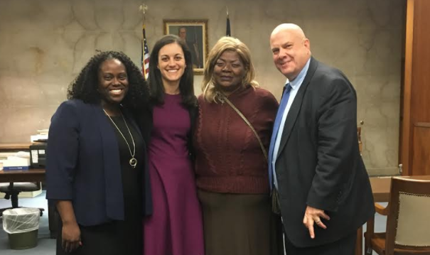 Four people stand together in a courtroom, smiling at the camera. The group consists of three women and one man dressed in formal attire. An American flag and framed picture are visible in the background.