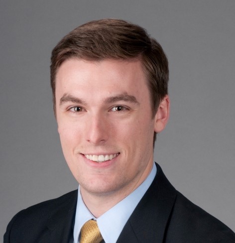 A person with short brown hair wearing a suit, light blue shirt, and yellow tie, smiles at the camera against a plain gray background.