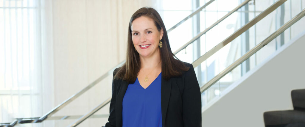 A woman with long brown hair, wearing a blue blouse and black blazer, stands smiling in front of a modern staircase with metal railings, set in a well-lit interior space.