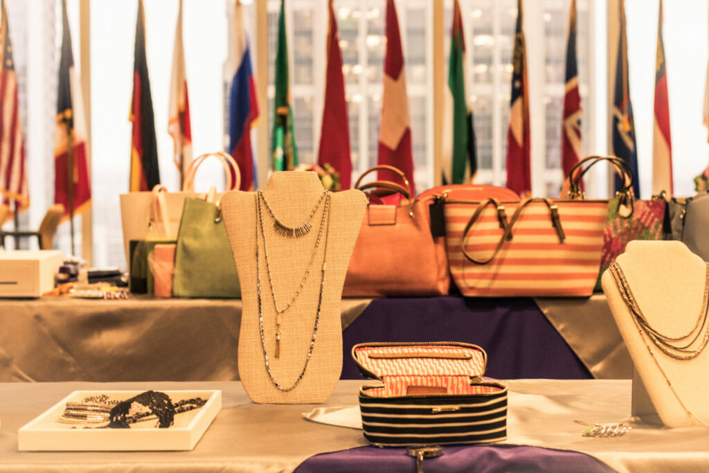 A display table featuring elegant necklaces on stands, bracelets in a box, and a variety of handbags in the background. Multiple international flags are visible, indicating a global theme or event.