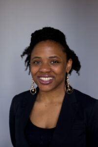 A person with curly hair is smiling while wearing a black blazer and statement earrings against a gray background.