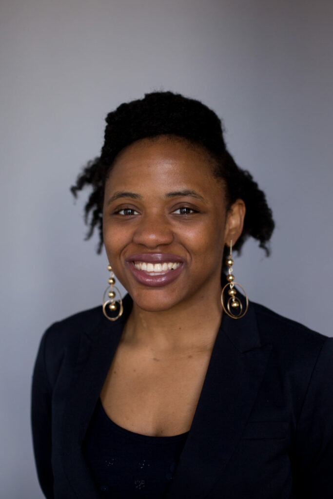 A person with curly hair is smiling while wearing a black blazer and statement earrings against a gray background.
