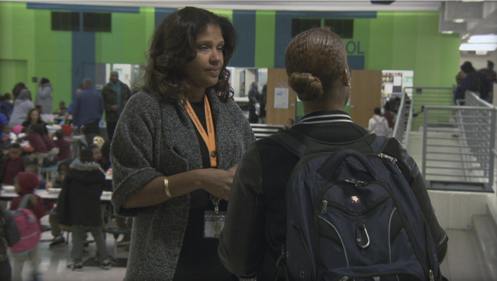 Two people are conversing in a busy indoor setting. One is facing the camera wearing a grey cardigan and lanyard, while the other has their back to the camera wearing a backpack. The background shows multiple people and green walls.