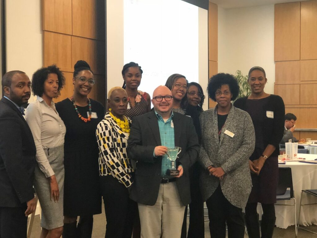 A group of people standing indoors, posing for a photo. One person at the center holds a trophy and smiles. They're in a conference room with a projector screen in the background. The group is diverse, with individuals dressed in business attire.