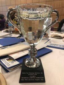 A trophy awarded to the Atlanta Volunteer Lawyers Foundation by Metro Fair Housing Services, Inc. for the 2018 Freedom Fighter Cup. The trophy is placed on a table surrounded by papers and folders.