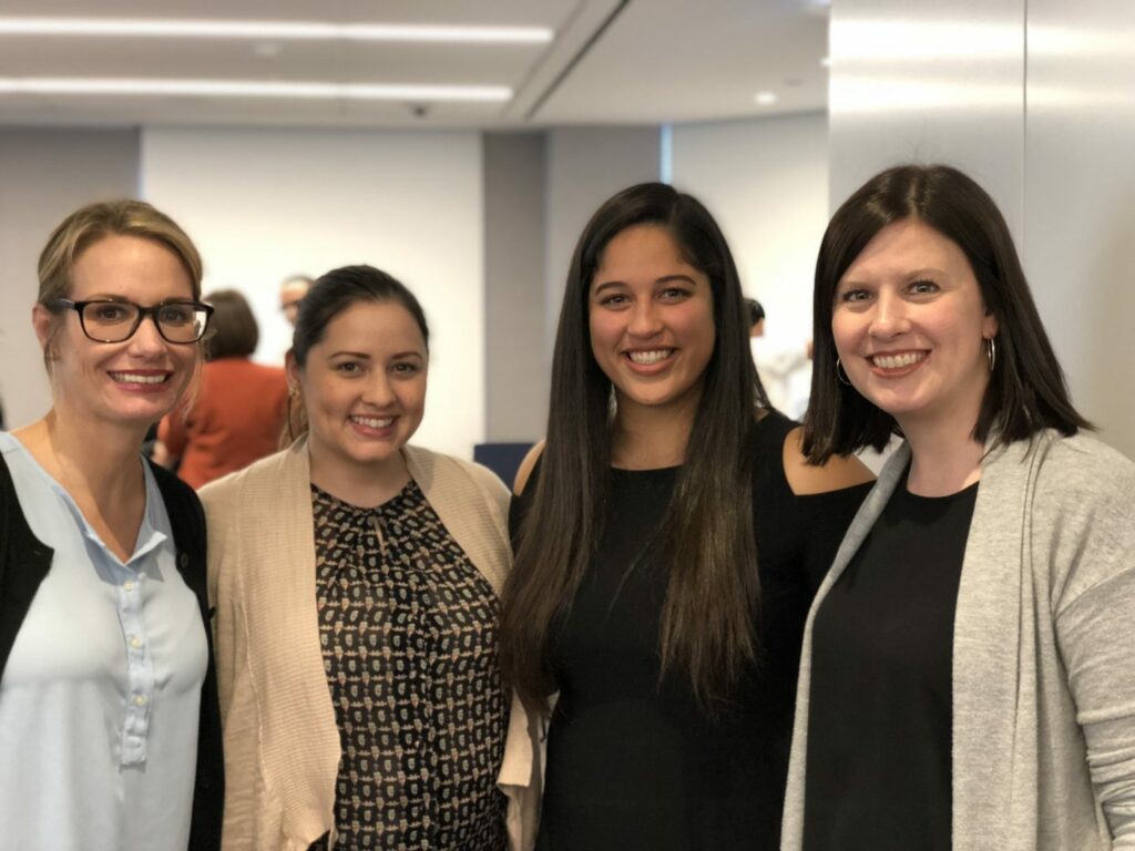 Four women smiling and posing together in an indoor setting with neutral-colored walls and ceiling lights in the background. They are wearing casual business attire.