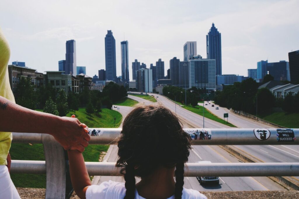 A child with braided hair stands on a bridge overlooking a highway leading to a city skyline, holding hands with an adult. Towers rise in the distance, and the scene is bathed in daylight.