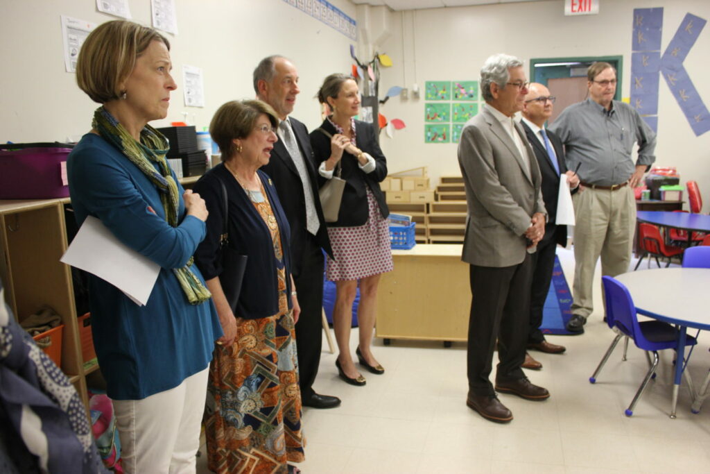 A group of seven adults stand in a classroom, wearing business and casual attire. Some are holding papers. The room has children's furniture and educational posters on the walls.